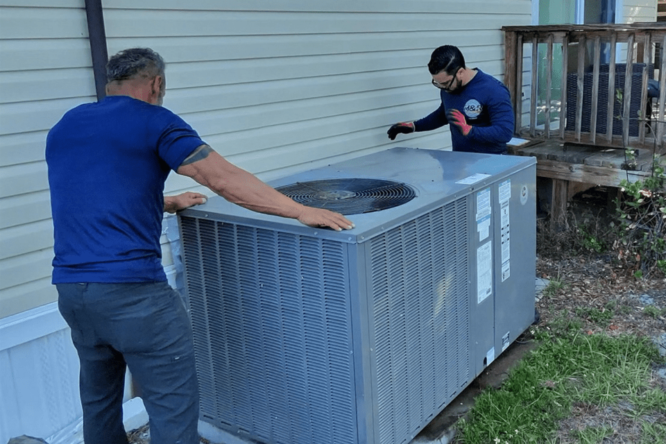 Two HVAC technicians installing a large residential AC unit outdoors, ensuring proper setup and safety, highlighting professional air conditioning services.